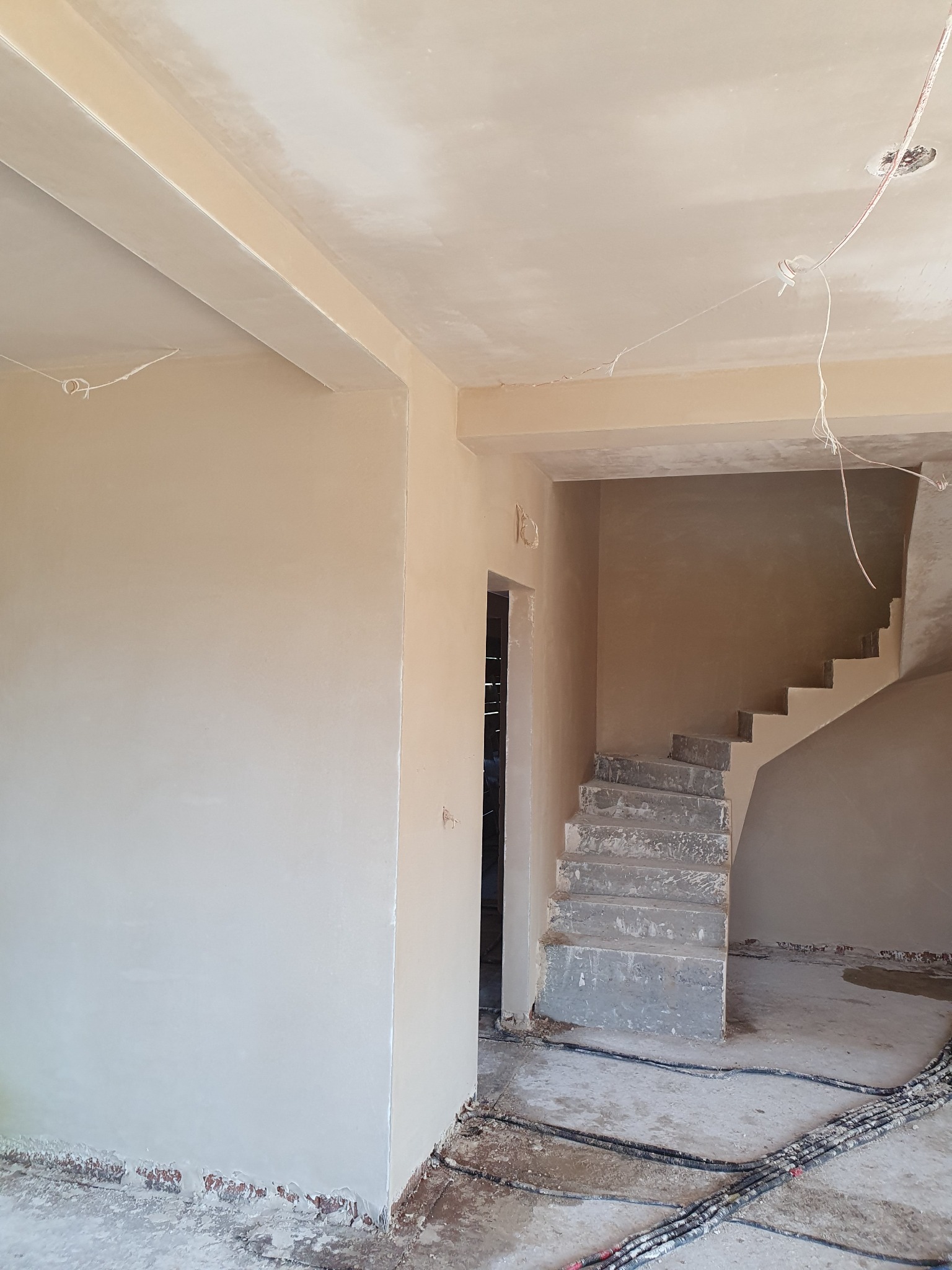 Wnętrze w trakcie prac wykończeniowych: świeżo otynkowane ściany i sufit w jasnym beżu, surowe betonowe schody w tle, widoczne kable elektryczne na podłodze.
