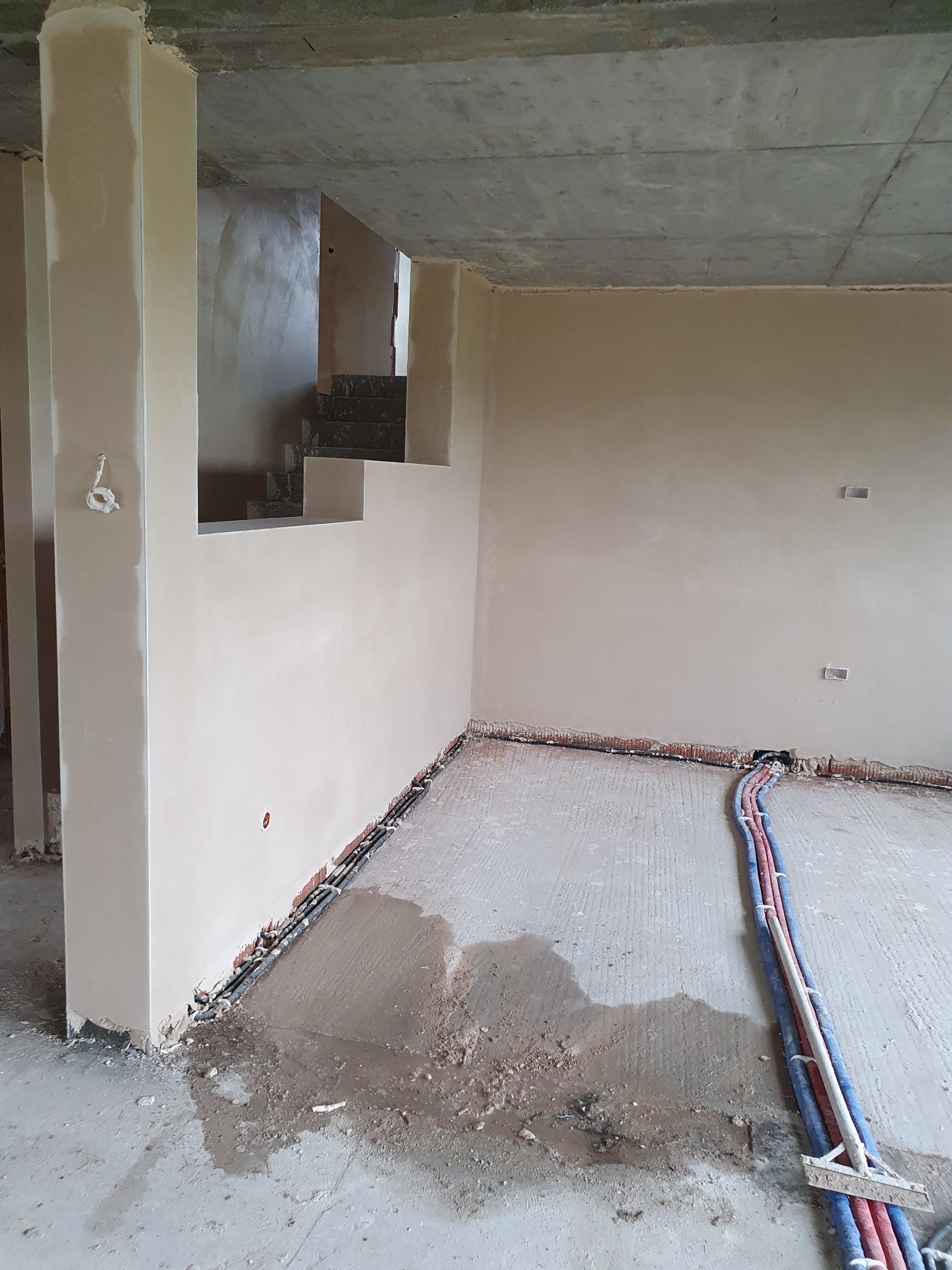 Surowe wnętrze z tynkami maszynowymi na ścianach, widoczne instalacje elektryczne i hydrauliczne wzdłuż podłogi, fragment schodów w tle.