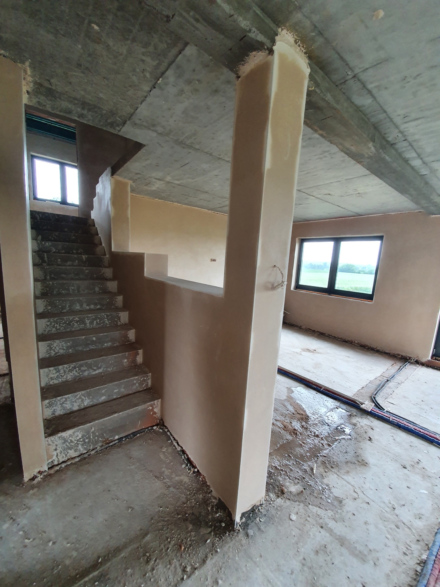 Wnętrze w stanie surowym po nałożeniu tynków. Widoczne schody, słup nośny i ściany pokryte świeżym tynkiem w odcieniach beżu. Na podłodze widoczne ślady wody i przewody elektryczne.
