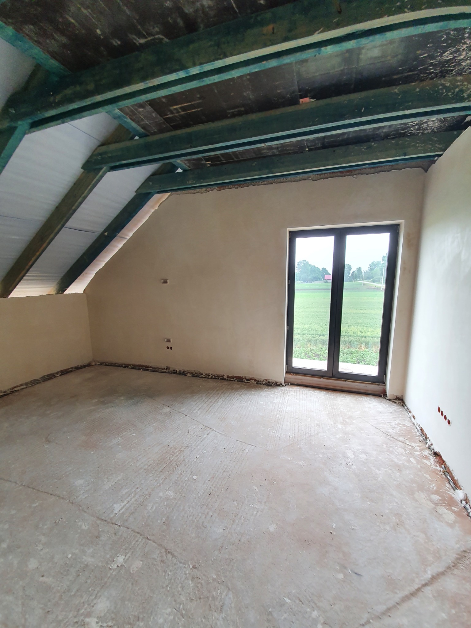 Poddasze po nałożeniu tynków, widoczne drewniane elementy konstrukcji dachu pomalowane na zielono, ściany w kolorze beżowym, okno z widokiem na pole.