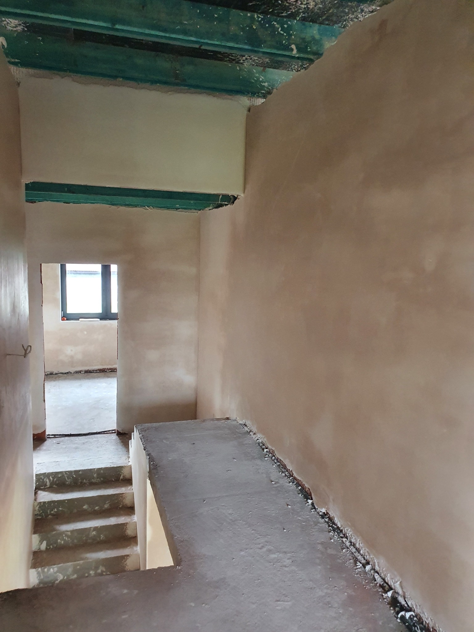 Wnętrze budynku w trakcie prac wykończeniowych: świeżo nałożone tynki na ścianach, widoczne schody i surowy strop z elementami konstrukcyjnymi pomalowanymi na zielono.
