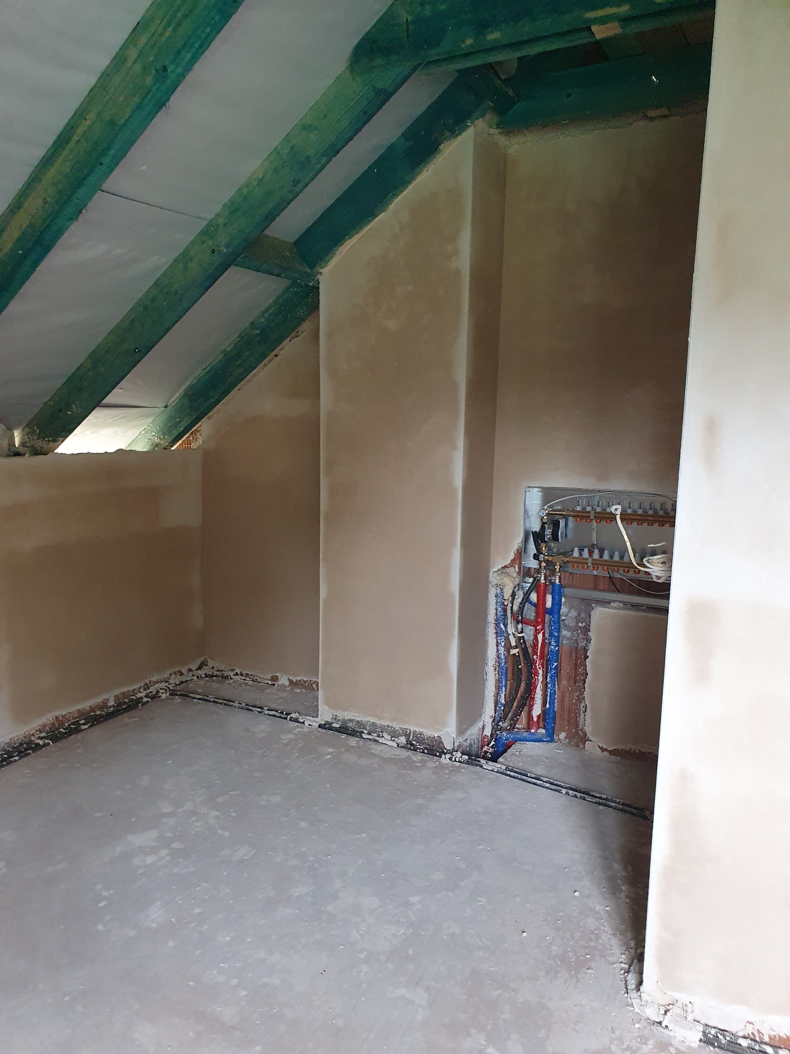 Pokój na poddaszu w trakcie prac wykończeniowych, ściany pokryte świeżym tynkiem, widoczne fragmenty instalacji grzewczej i zielone belki stropowe.