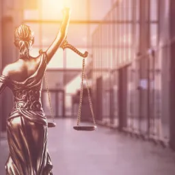 Złota statuetka Temidy, bogini sprawiedliwości z wagą i mieczem, widziana od tyłu na tle nowoczesnego, przeszklonego budynku biurowego.