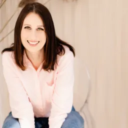 Portret uśmiechniętej kobiety w różowej koszuli, siedzącej na przezroczystym krześle na tle beżowej ściany z dekoracjami z suszonych traw.