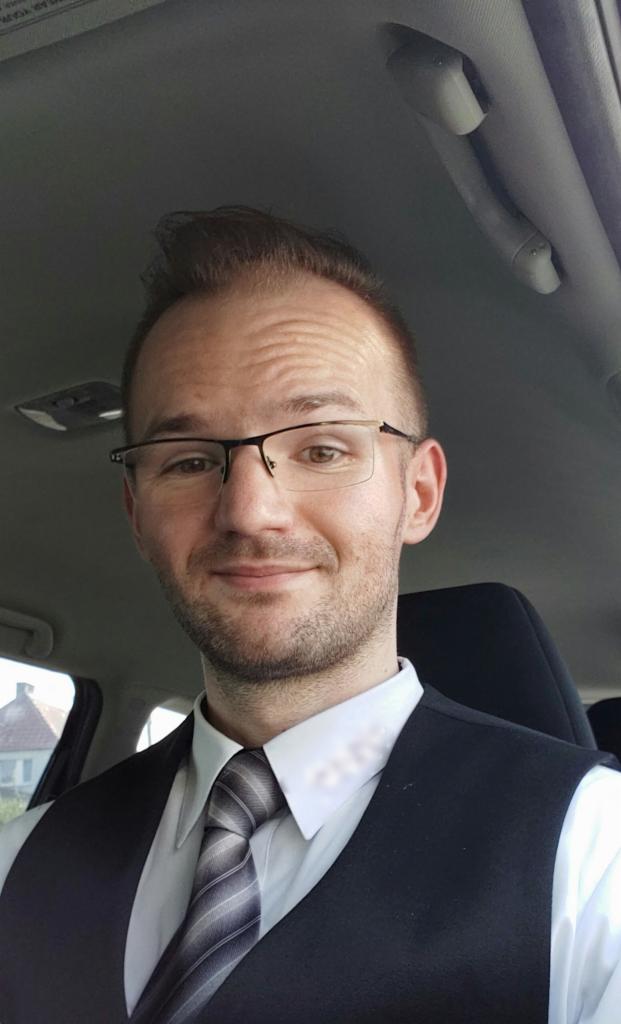 Portret mężczyzny w okularach, ubrany w koszulę, krawat i kamizelkę, siedzącego w samochodzie. Uśmiech na twarzy, jasne oświetlenie wnętrza pojazdu.