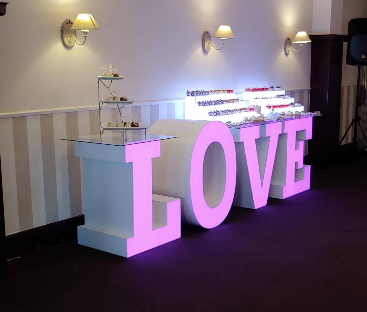 Podświetlany napis LOVE z deserami na stole i trzypoziomowym stojaku na słodkości, dekoracja sali weselnej z kinkietami na ścianie.