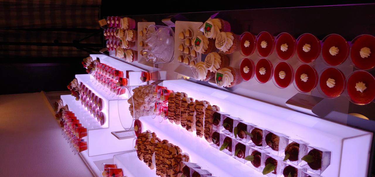 Elegancki, podświetlany bufet z różnorodnymi deserami: mini galaretki, babeczki z logo, kremowe desery w szklankach, ciasteczka i czekoladowe kulki z listkiem mięty.