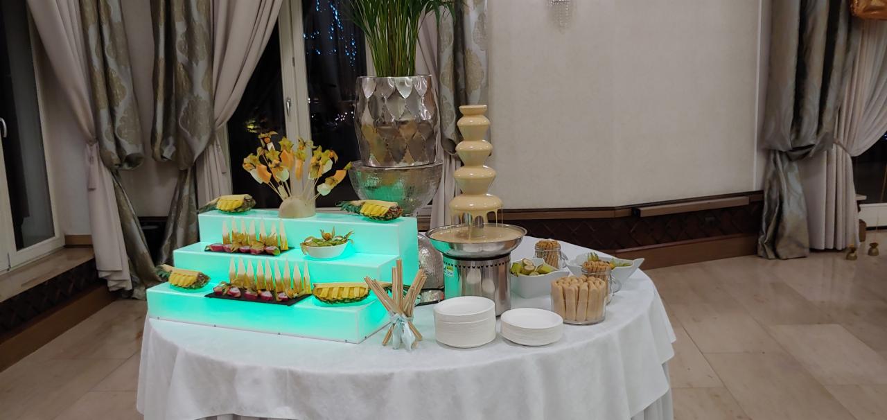 Elegancki stół z fontanną białej czekolady, owocami, ciasteczkami i przekąskami na podświetlanym, trójstopniowym podeście, udekorowany kwiatami i roślinami w srebrnych wazonach, widok z przodu.