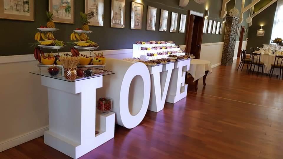 Stół z przekąskami owocowymi i deserami na przyjęciu, ustawiony przed dużym napisem LOVE z białych liter, drewniana podłoga, eleganckie dekoracje sali.