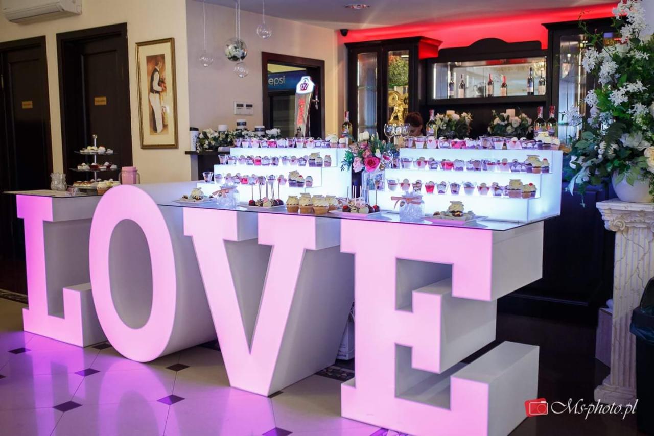 Dekoracyjny stół z podświetlanymi literami LOVE, na którym ustawione są desery i napoje w małych porcjach, w tle bufet z alkoholem i kwiatami.