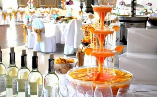 Fontanna z pomarańczowego napoju z plasterkami pomarańczy na stole z butelkami wina i przekąskami, przygotowana na przyjęcie w jasnej sali.