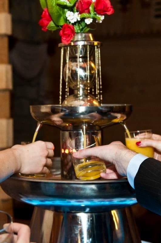 Fontanna z napojem pomarańczowym, dekorowana kwiatami, nalewająca napój do szklanek trzymanych przez osoby w garniturze i z czerwonym lakierem na paznokciach.