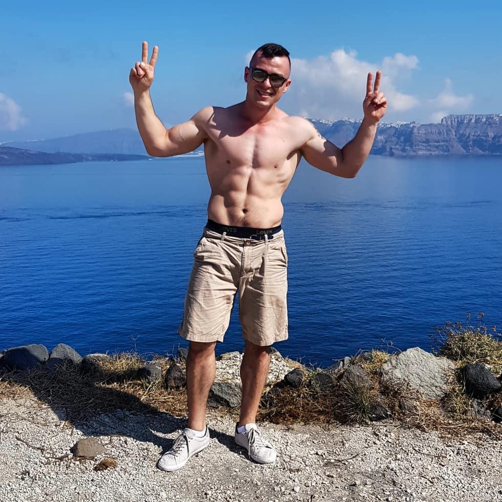 Uśmiechnięty, opalony mężczyzna prezentuje wyrzeźbioną sylwetkę na tle błękitnego morza i gór, pozdrawiając dwoma palcami. Okulary przeciwsłoneczne i białe buty.