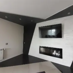 Nowoczesny salon z białą ścianą wyłożoną dekoracyjną cegłą, na której wisi telewizor i czarny, podłużny kominek elektryczny, z czarno-białą podłogą w szachownicę i białym grzejnikiem.