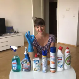 Uśmiechnięta kobieta w bluzce w panterkę i niebieskich rękawicach, prezentująca gest OK obok zestawu środków czystości różnych marek na białym okrągłym stole w jasnym wnętrzu.