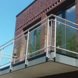Szklana balustrada balkonowa z metalowymi elementami mocującymi, widoczna na tle ceglanej ściany budynku i błękitnego nieba.