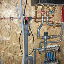 Rozdzielacz ogrzewania podłogowego z niebieskimi rurami, zamontowany na ścianie z płyty OSB, widoczne liczne zawory i rury miedziane.