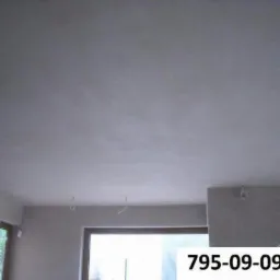 Gładka, jasnoszara powierzchnia sufitu po nałożeniu tynku, widoczne kable wystające z sufitu, w tle okno i fragment ściany z numerem telefonu.