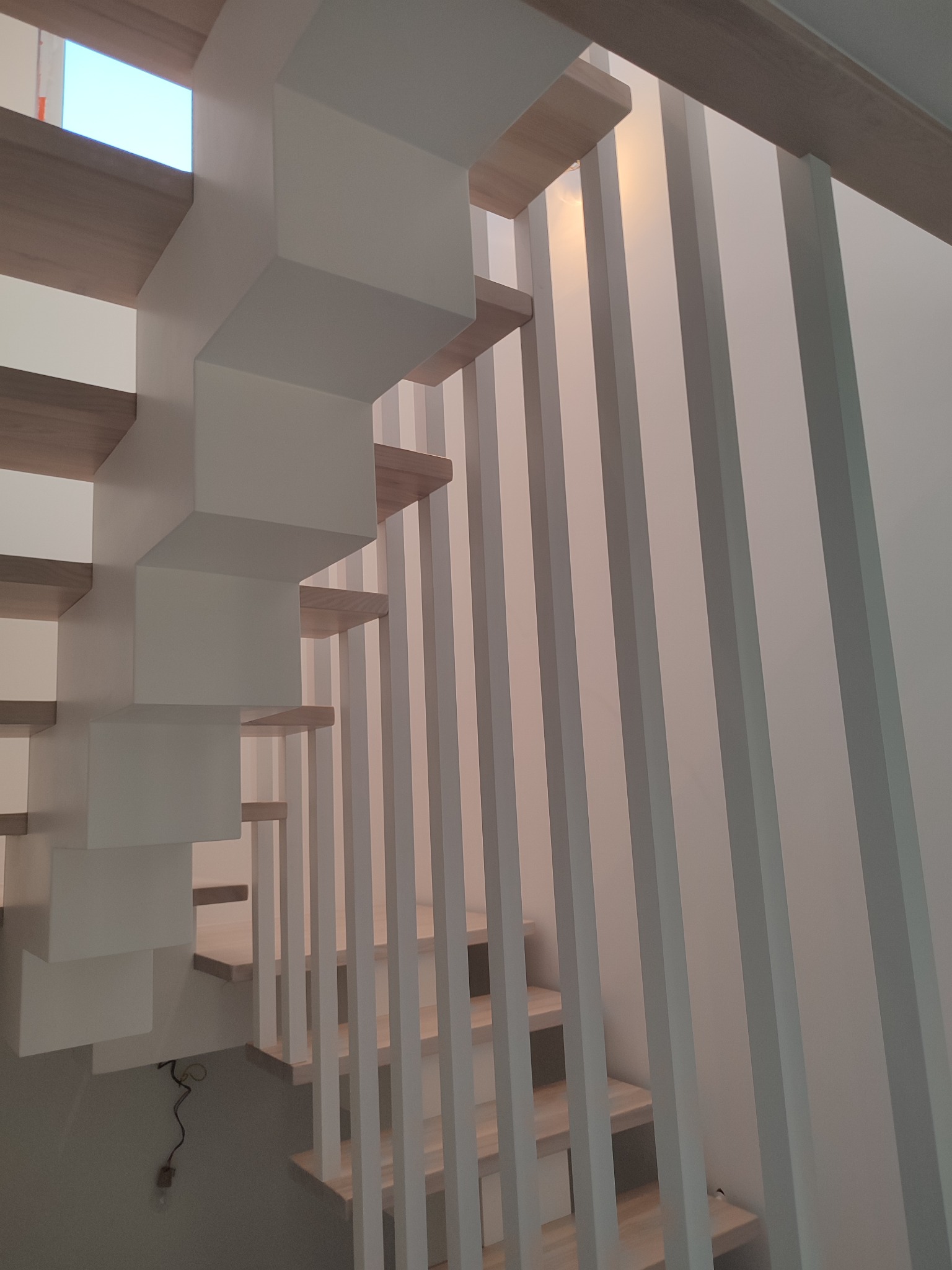 Nowoczesne schody z drewnianymi stopniami i białymi, geometrycznymi wspornikami oraz pionowymi balustradami, widziane z dołu, z widocznym fragmentem instalacji elektrycznej.