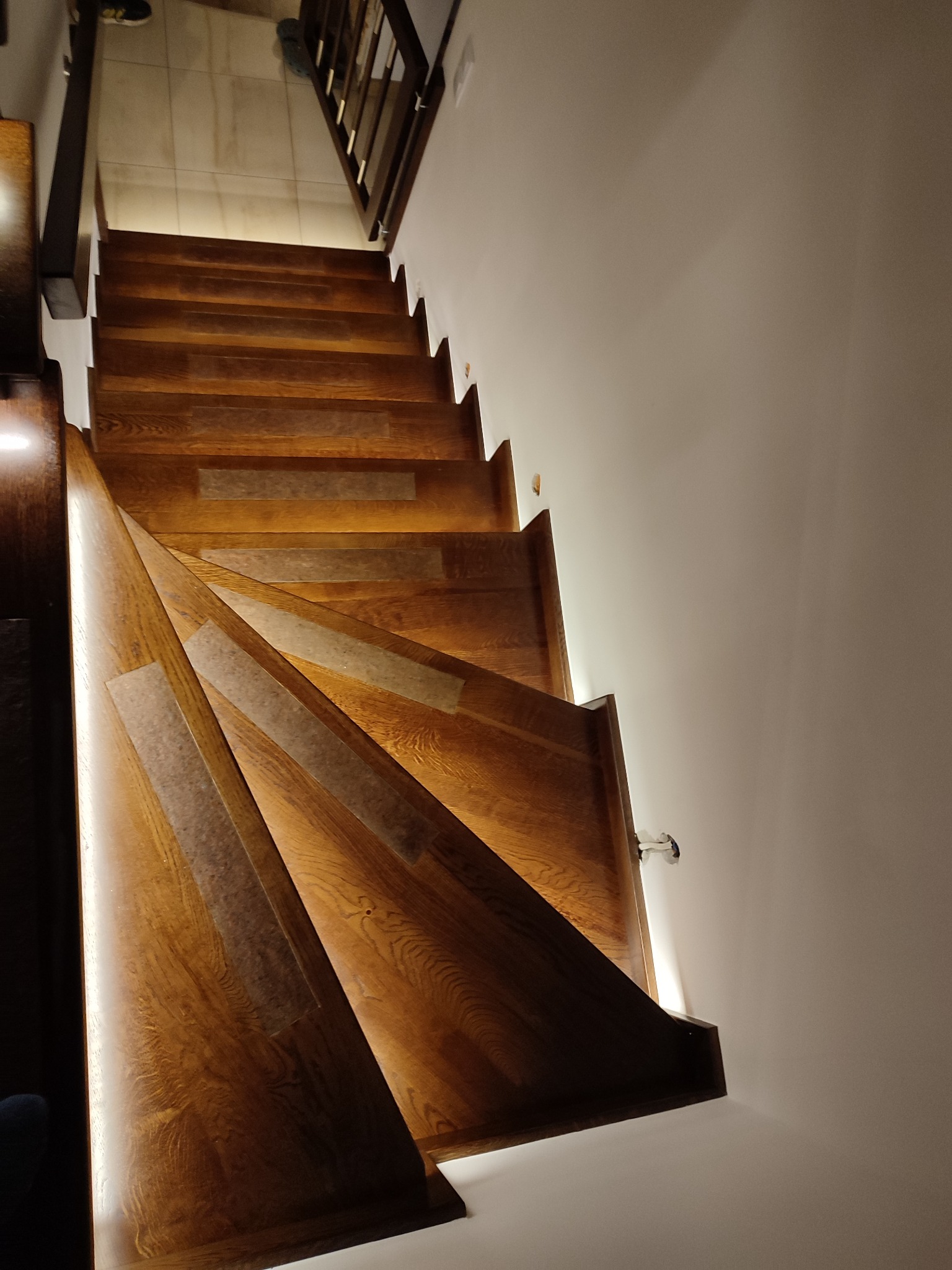 Drewniane schody z oświetleniem LED pod stopniami, widok z dołu, z widoczną fakturą drewna i skrętem biegów.