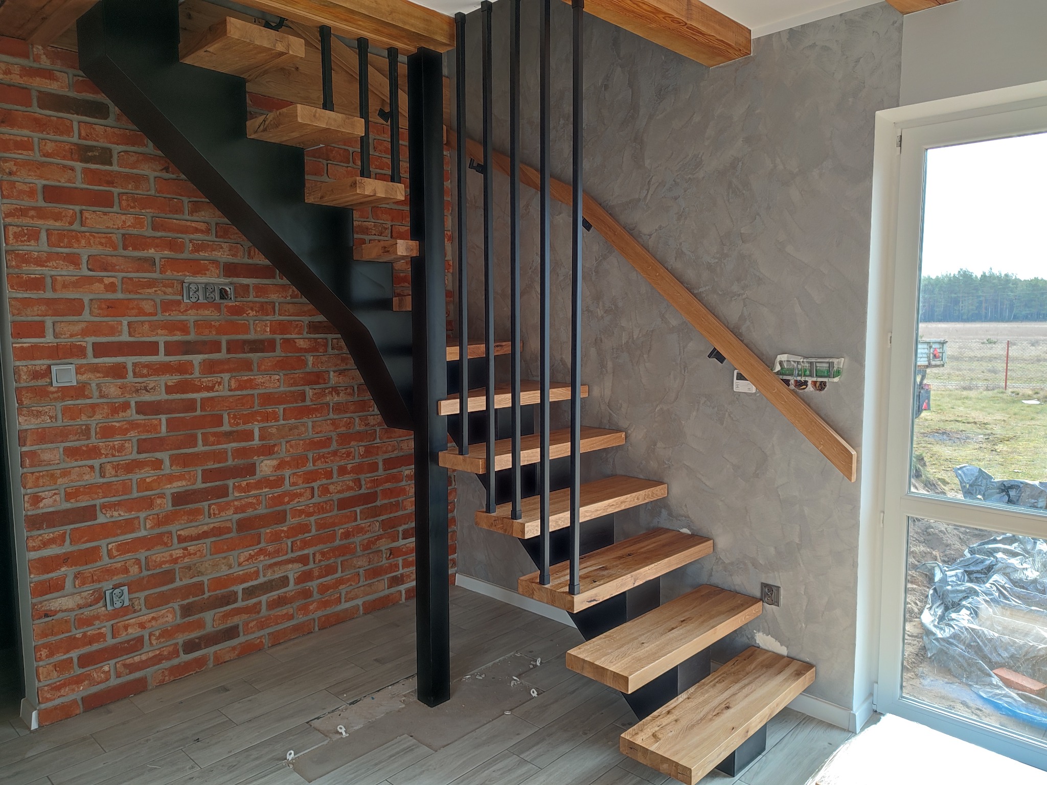 Nowoczesne schody z drewnianymi stopniami i czarną metalową konstrukcją, umieszczone w pomieszczeniu z ceglaną ścianą i szarą ścianą z efektem betonu.