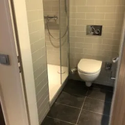 Wykończona łazienka z kabiną prysznicową i podwieszaną toaletą, szare płytki na ścianach i czarne na podłodze, widoczne detale takie jak przycisk spłuczki i uchwyt na papier toaletowy.