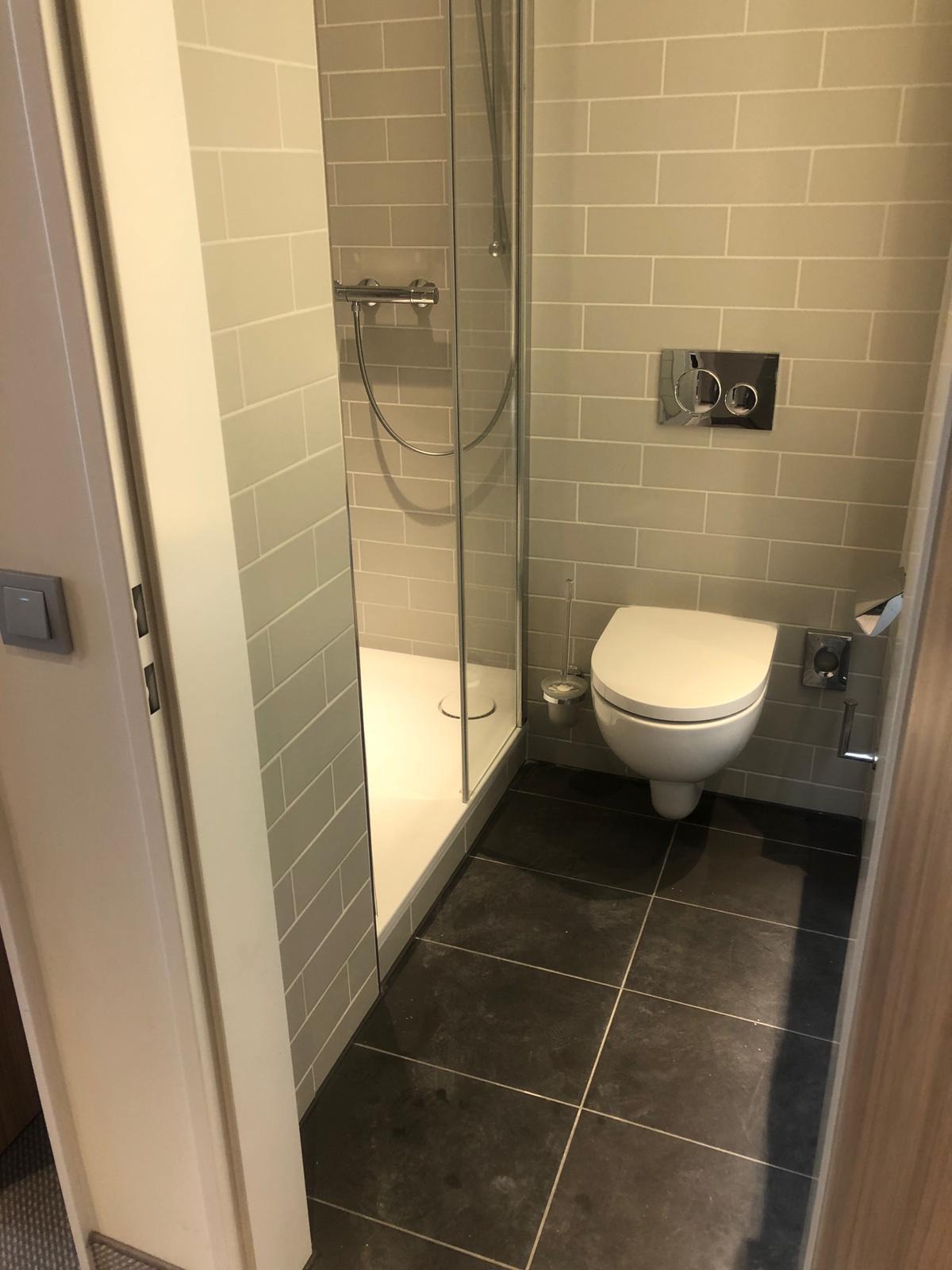 Wykończona łazienka z kabiną prysznicową i podwieszaną toaletą, szare płytki na ścianach i czarne na podłodze, widoczne detale takie jak przycisk spłuczki i uchwyt na papier toaletowy.