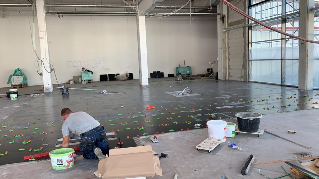 Układanie ciemnoszarych płytek podłogowych w dużym, surowym wnętrzu. Widoczne krzyżyki dystansowe, poziomica, wiaderka z klejem oraz glazurnik w trakcie pracy.