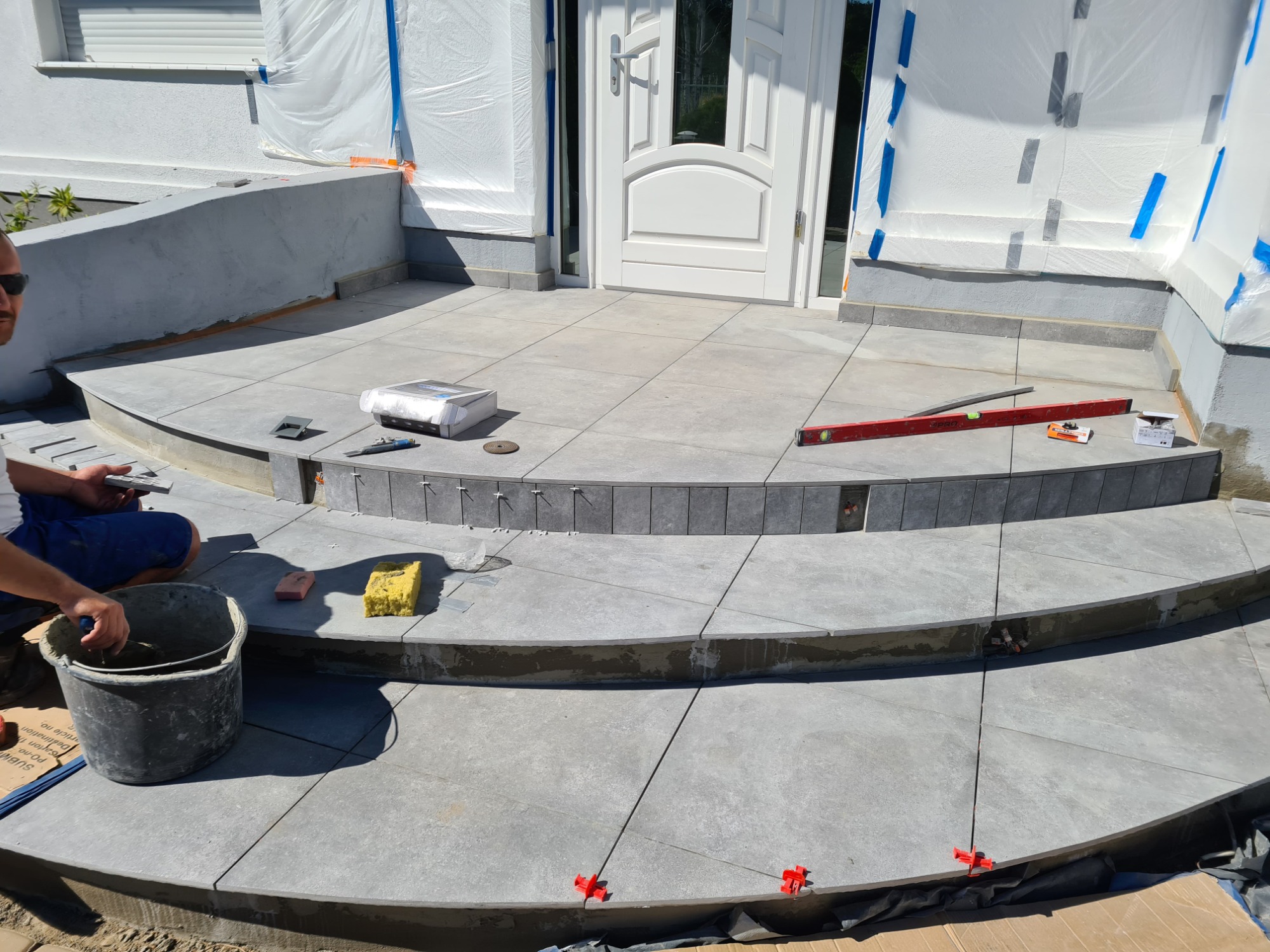 Układanie szarych płytek na schodach wejściowych w Starym Kisielinie, widoczne narzędzia glazurnicze, poziomica i kubeł z zaprawą.