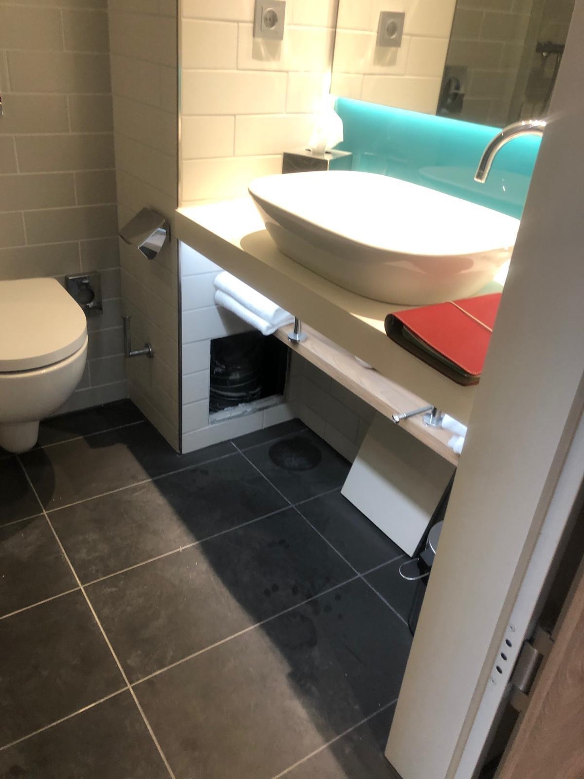 Nowoczesna łazienka z owalną umywalką nablatową, turkusowym panelem nad umywalką, szarą glazurą ścienną i ciemną, kwadratową podłogą z fugami oraz toaletą.