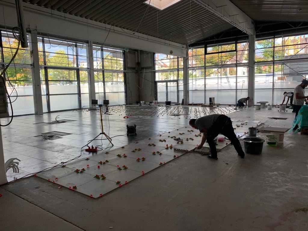 Układanie płytek podłogowych w dużej hali, widoczni pracownicy w trakcie pracy, używane poziomice i system poziomowania płytek, surowy stan pomieszczenia z widocznymi instalacjami.