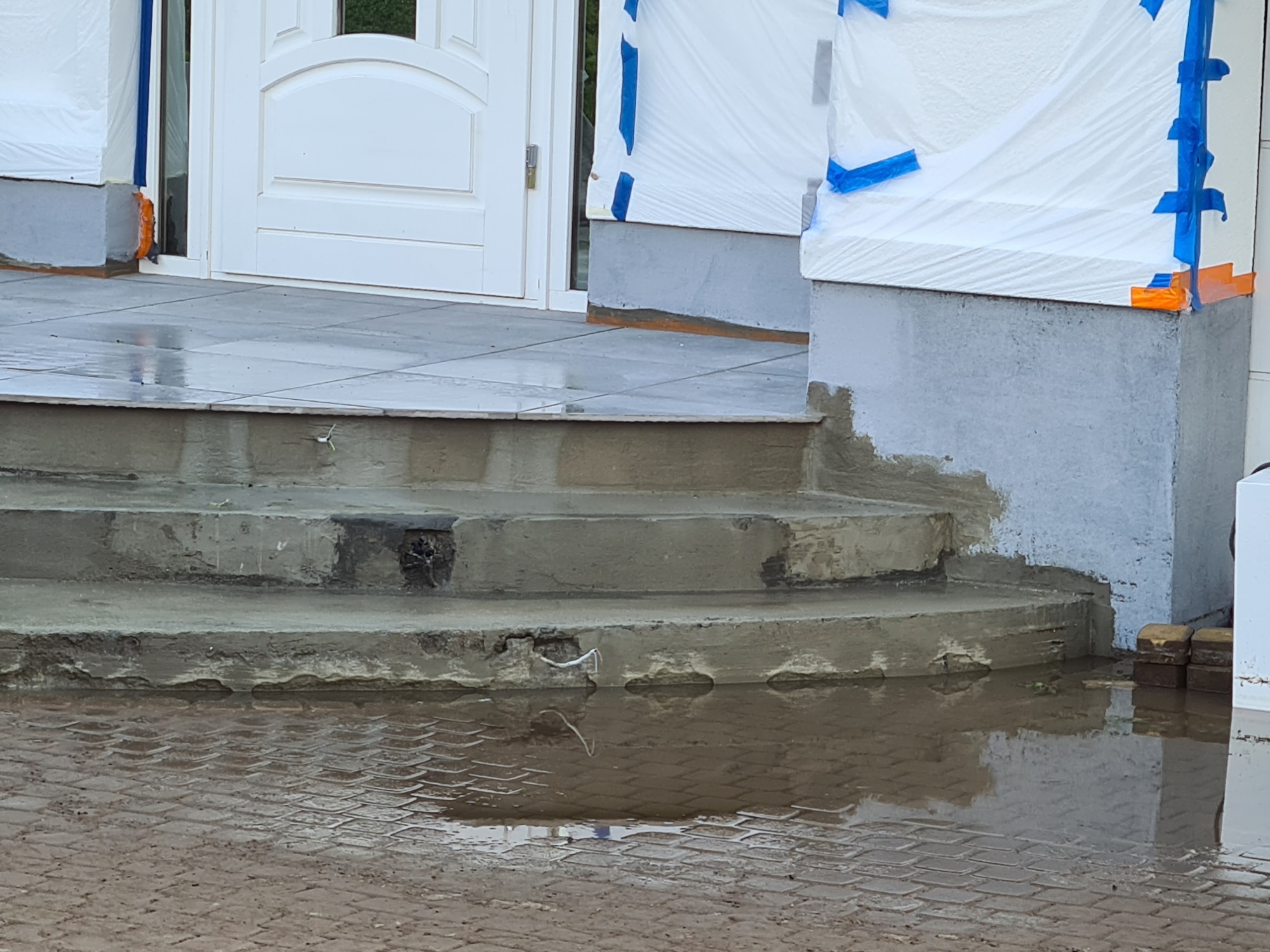 Wykończenie schodów wejściowych do domu z szarych płytek, widoczne ślady świeżego cementu i zabezpieczenia elewacji folią i taśmą malarską, kałuża na brukowanym chodniku odbija schody.