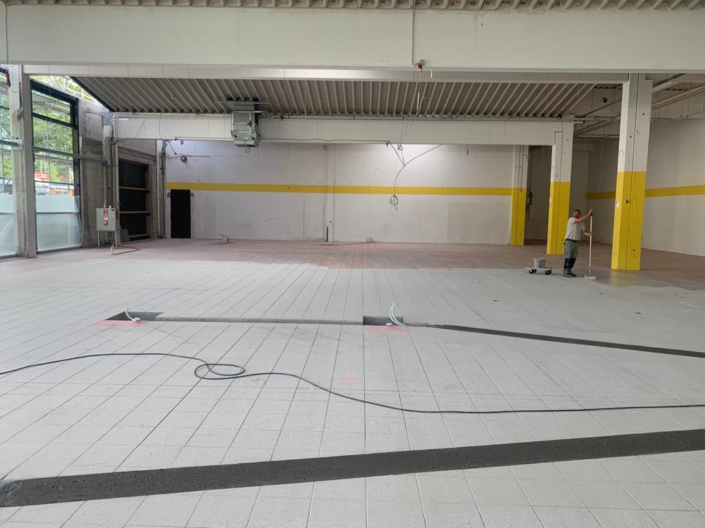 Wnętrze hali w trakcie malowania: białe ściany z żółtym pasem, szara podłoga z widocznymi kablami i szczelinami, malarz z wałkiem przy kolumnie z żółtą dolną częścią.