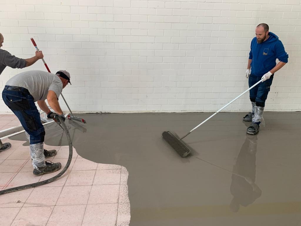 Ekipa budowlana w trakcie wylewania samopoziomującej wylewki na podłogę w pomieszczeniu z białymi ceglanymi ścianami. Widoczny fragment starej, różowej podłogi z płytek.