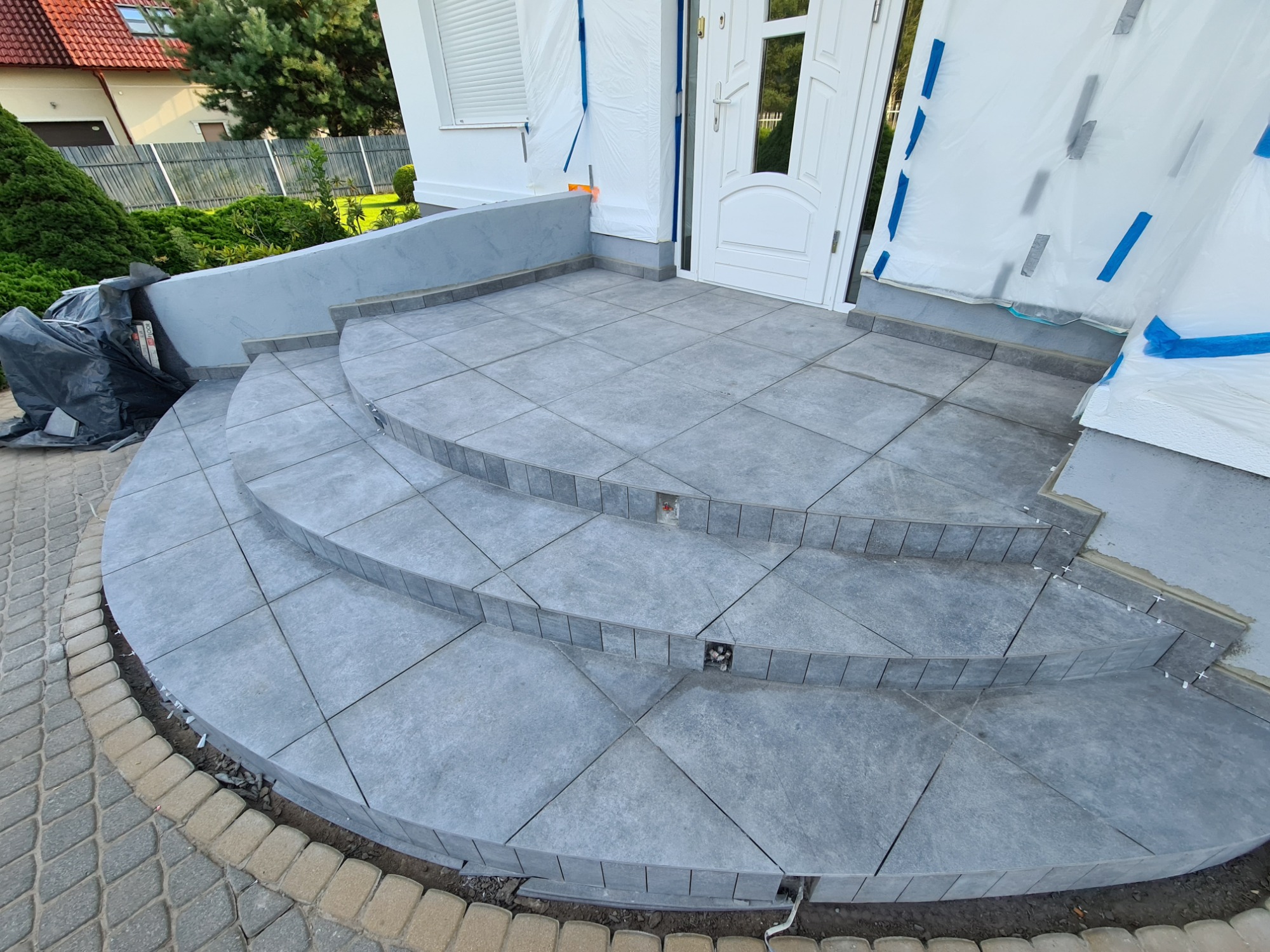 Wyłożone szarymi płytkami schody wejściowe do domu, z widocznymi krzyżykami dystansowymi między płytkami, zabezpieczone folią okna i drzwi.