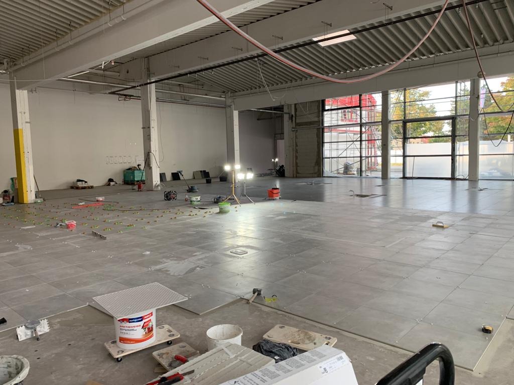 Układanie szarych płytek podłogowych w przestronnym, surowym wnętrzu hali; widoczne narzędzia glazurnicze, poziomica, krzyżyki dystansowe, wiadro z klejem na wózku i lampy oświetlające obszar roboczy.