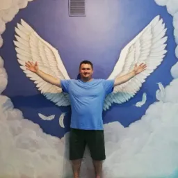 Mężczyzna w niebieskiej koszulce stoi na tle ściany z namalowanymi anielskimi skrzydłami i chmurami, rozkładając ręce.