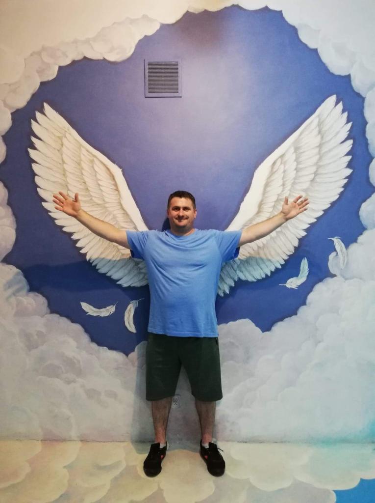Mężczyzna w niebieskiej koszulce stoi na tle ściany z namalowanymi anielskimi skrzydłami i chmurami, rozkładając ręce.