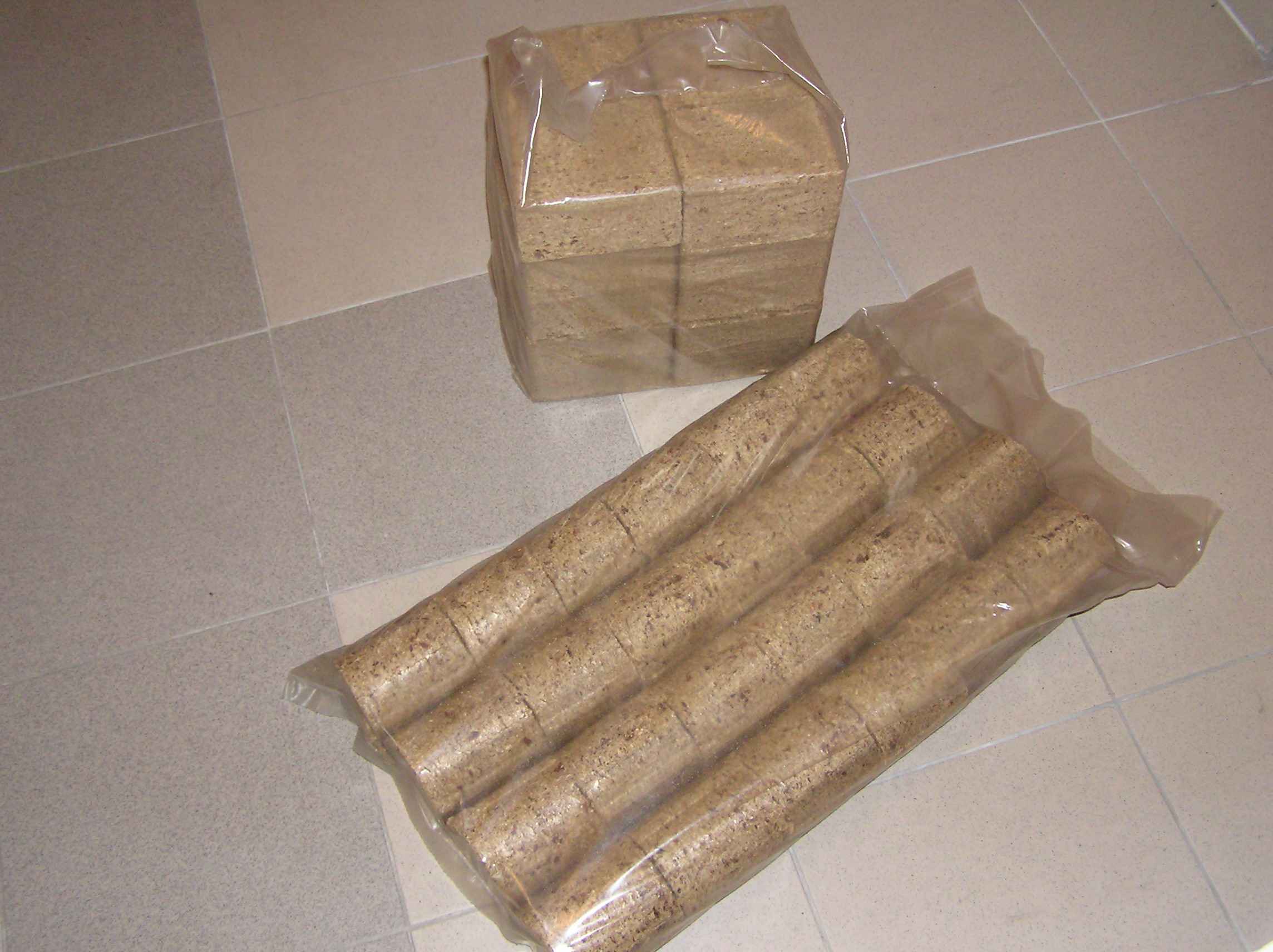 Dwa opakowania brykietu drzewnego: jedno w kostkach, drugie w wałkach, oba zapakowane w przezroczystą folię, leżą na kafelkach podłogowych.