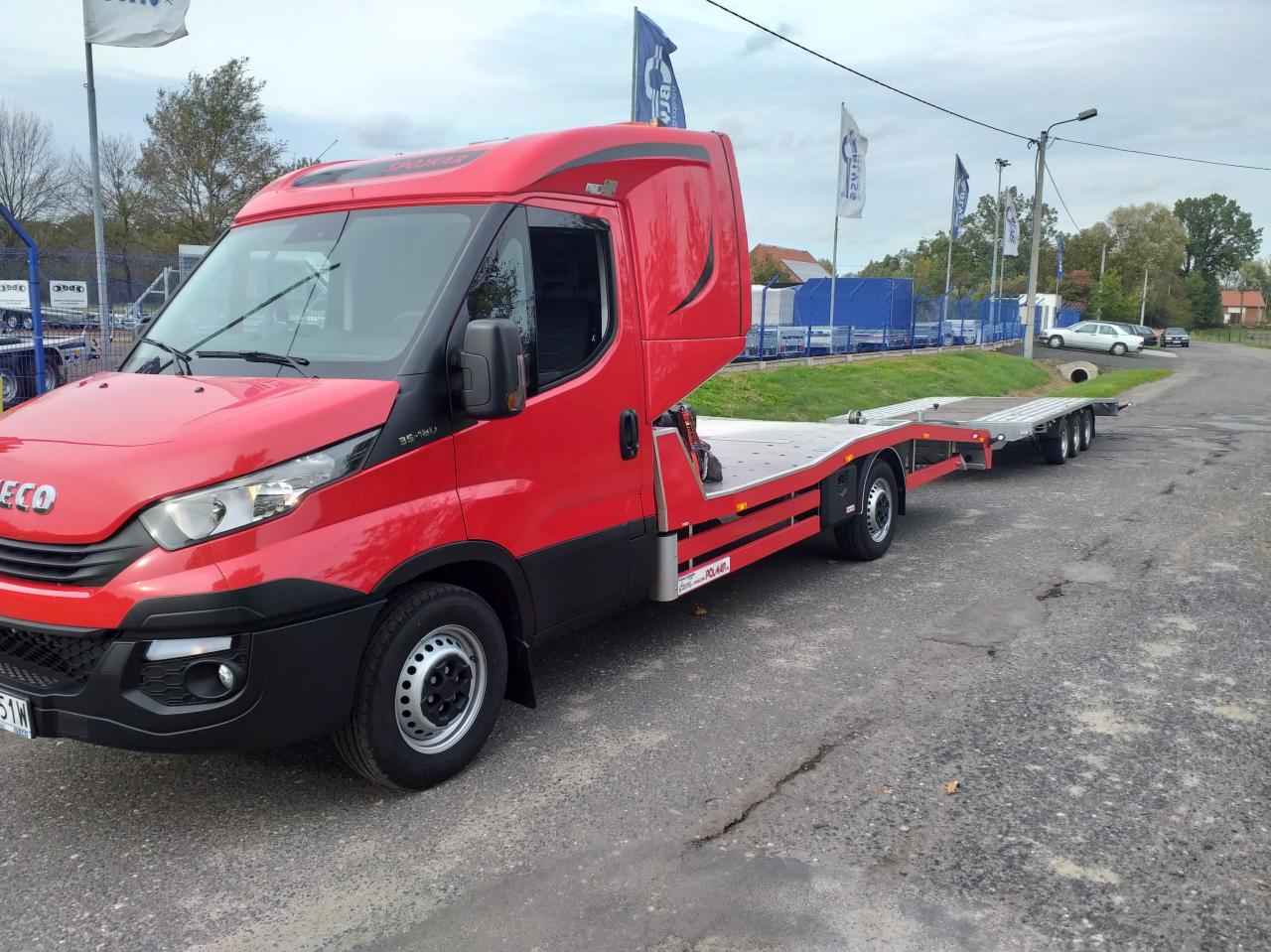 Czerwona laweta Iveco na szarej asfaltowej drodze, widok z boku na tle zieleni i budynków.