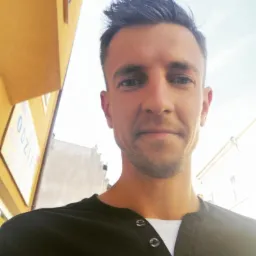 Portret mężczyzny z krótką fryzurą, ubranego w czarną koszulę z guzikami, na tle budynków w słoneczny dzień. Ujęcie z dołu.