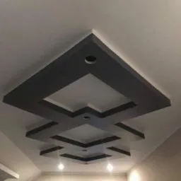 Nowoczesny sufit z geometrycznym wzorem z płyt kartonowo-gipsowych, podświetlony punktowymi lampami, w jasnym wnętrzu.