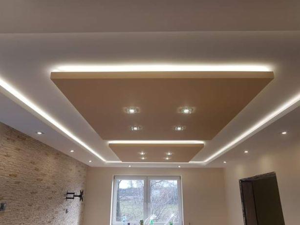 Nowoczesny sufit podwieszany z oświetleniem LED w kształcie kwadratów, jasne wnętrze z oknem i ceglaną ścianą w tle. Dekoracyjne wykończenie.