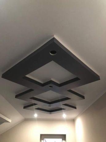 Nowoczesny sufit z geometrycznym wzorem z płyt kartonowo-gipsowych, podświetlony punktowymi lampami, w jasnym wnętrzu.