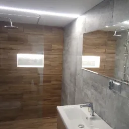 Nowoczesna łazienka z kabiną prysznicową typu walk-in, wykończona płytkami imitującymi drewno i szarością, oświetlenie LED podsufitowe, umywalka nablatowa z szafką w kolorze drewna.