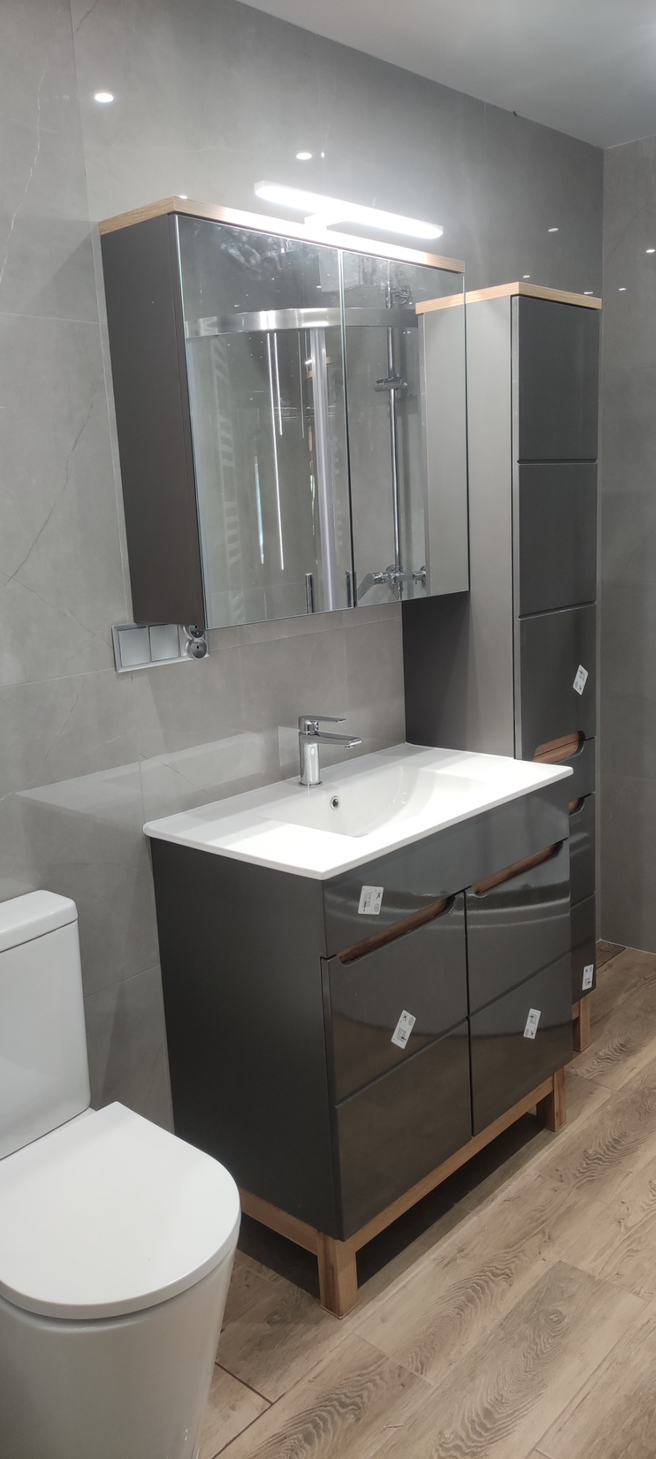Kompletna aranżacja łazienki: szara umywalka z szafką, lustro z oświetleniem, słupek i toaleta, na tle szarych płytek i drewnopodobnej podłogi.