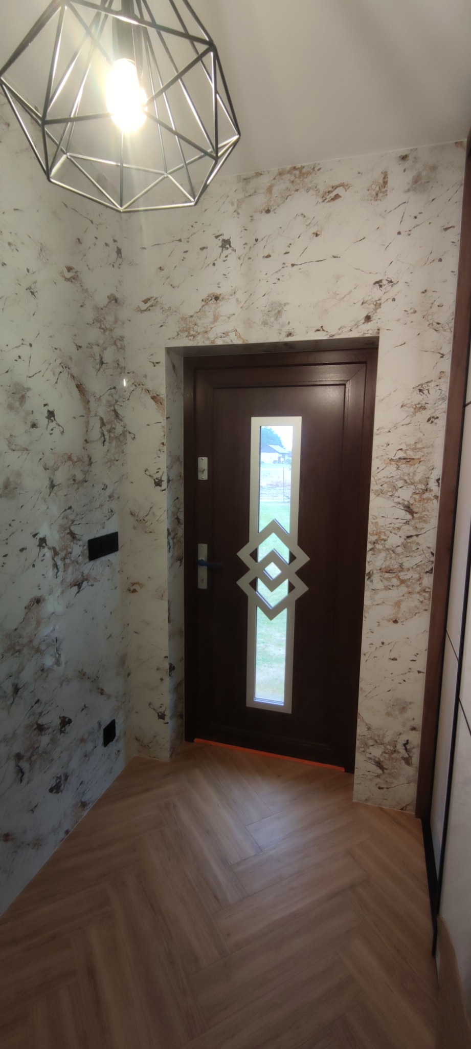 Przedpokój z podłogą ułożoną w jodełkę, ściany wykończone efektem marmuru, brązowe drzwi z szybą o geometrycznym wzorze i nowoczesna lampa sufitowa.