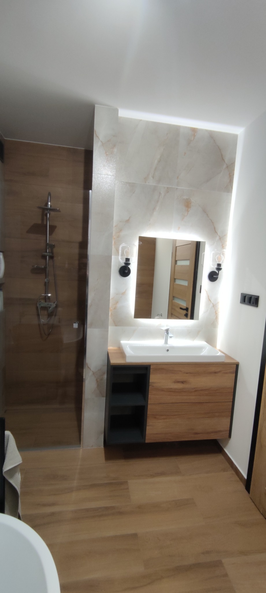 Nowoczesna łazienka z drewnopodobną podłogą i ścianami, kabina prysznicowa ze szklaną ścianą, umywalka z szafką w odcieniach drewna i szarości, lustro podświetlone od tyłu, kinkiety w stylu...