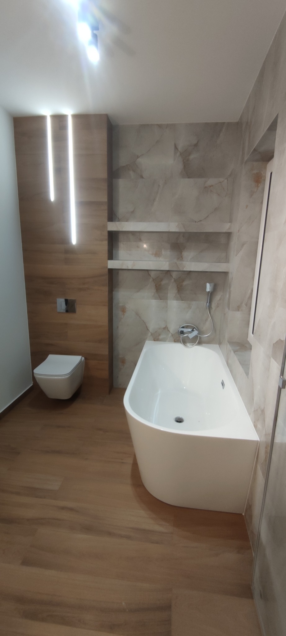 Nowoczesna łazienka z wanną wolnostojącą, podświetlaną ścianą z toaletą podwieszaną, półkami w ścianie wyłożonej płytkami imitującymi marmur oraz drewnopodobną podłogą.