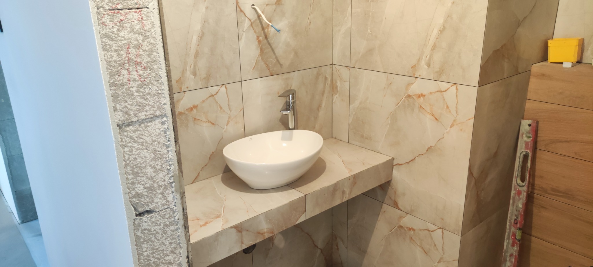 Niedokończona łazienka z owalną umywalką na blacie z płytek imitujących marmur, widoczne płytki na ścianach, wystające kable i poziomica oparta o drewnianą okładzinę.